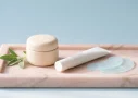 Tretinoin under eyes, with a porcelain cream pot, white tube and cooling gel pads on a pale pink marble tray